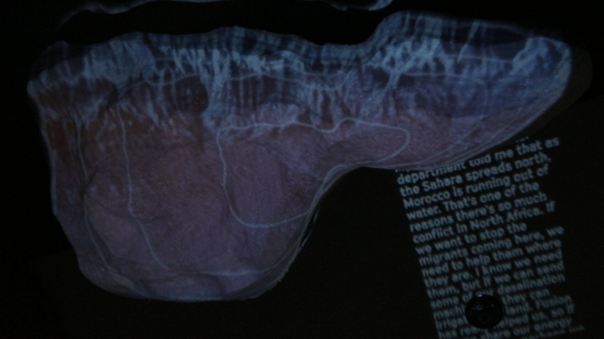 A topographic data-slice of North Africa is projected onto a curved surface, overlaid with scrolling policy narratives about water scarcity and migrationāone of the three synchronized āmission slicesā that anchor each loopās storyline.